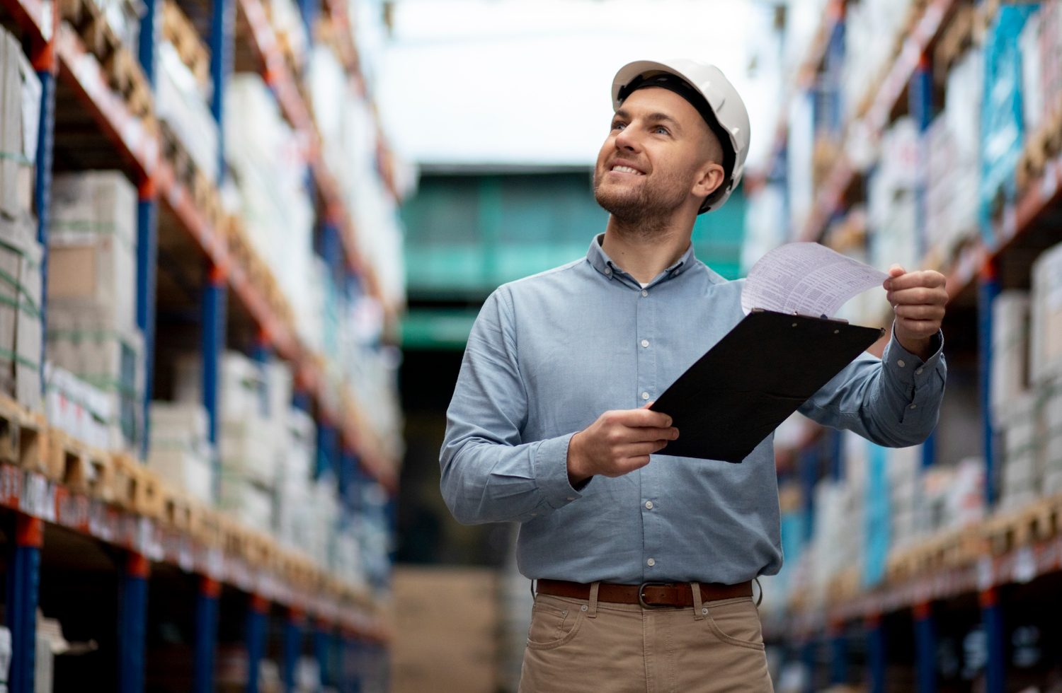 Male warehouse worker wearing a white hard hat and blue shirt, holding a clipboard and inspecting shelves.