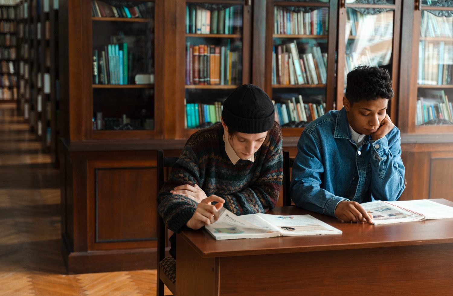 Two students sit at a wooden table in a library, reading open books and magazines side by side.
