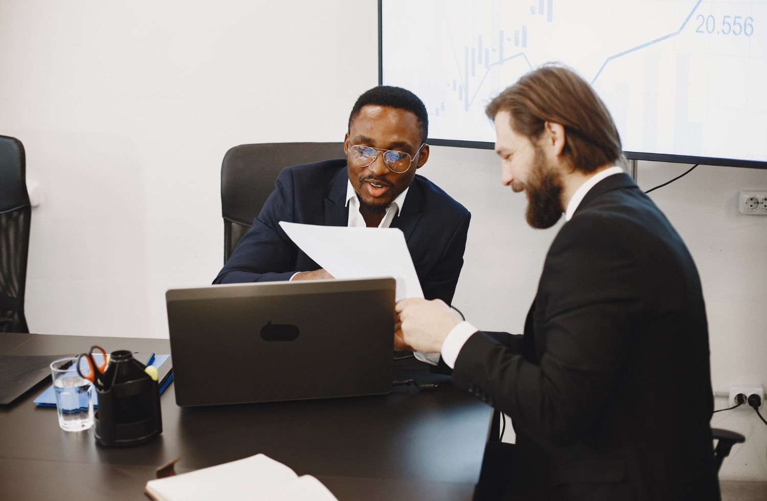 Two suited men discuss documents across a conference table, one showing papers while a laptop rests nearby, with a charted screen in the background