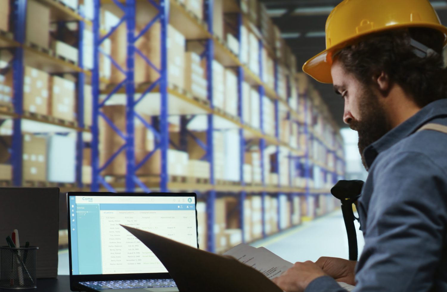 Warehouse worker in a yellow hard hat and blue uniform reviewing a document at a laptop in a warehouse aisle filled with tall shelves.
